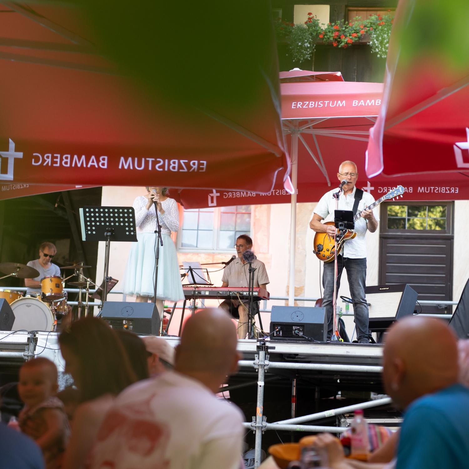 Bühne mit den Musikerinnen und Musikern, im Vordergrund unscharf Menschen an Biertischen